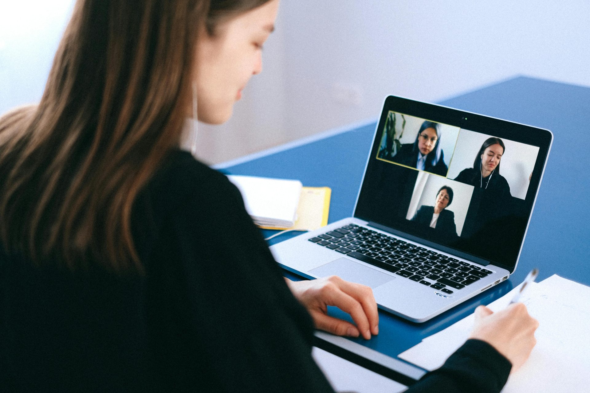 A woman looks towards her notepad and writes notes as she is on a call on her laptop with three othes