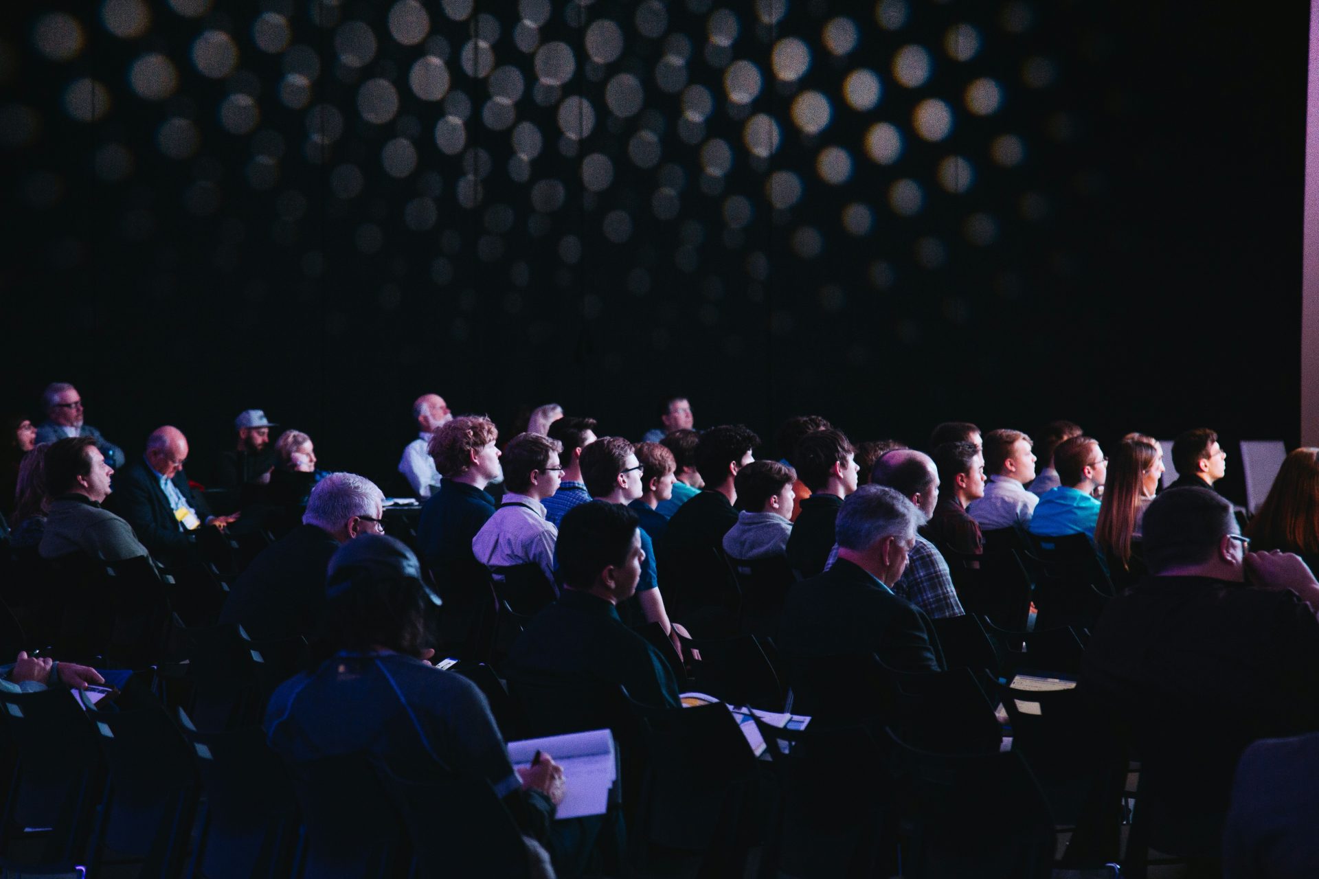 headway-F2KRf_QfCqw-unsplash A dark, wide shot of an audience sat down, facing a speaker with light glaring at them and all listening intently