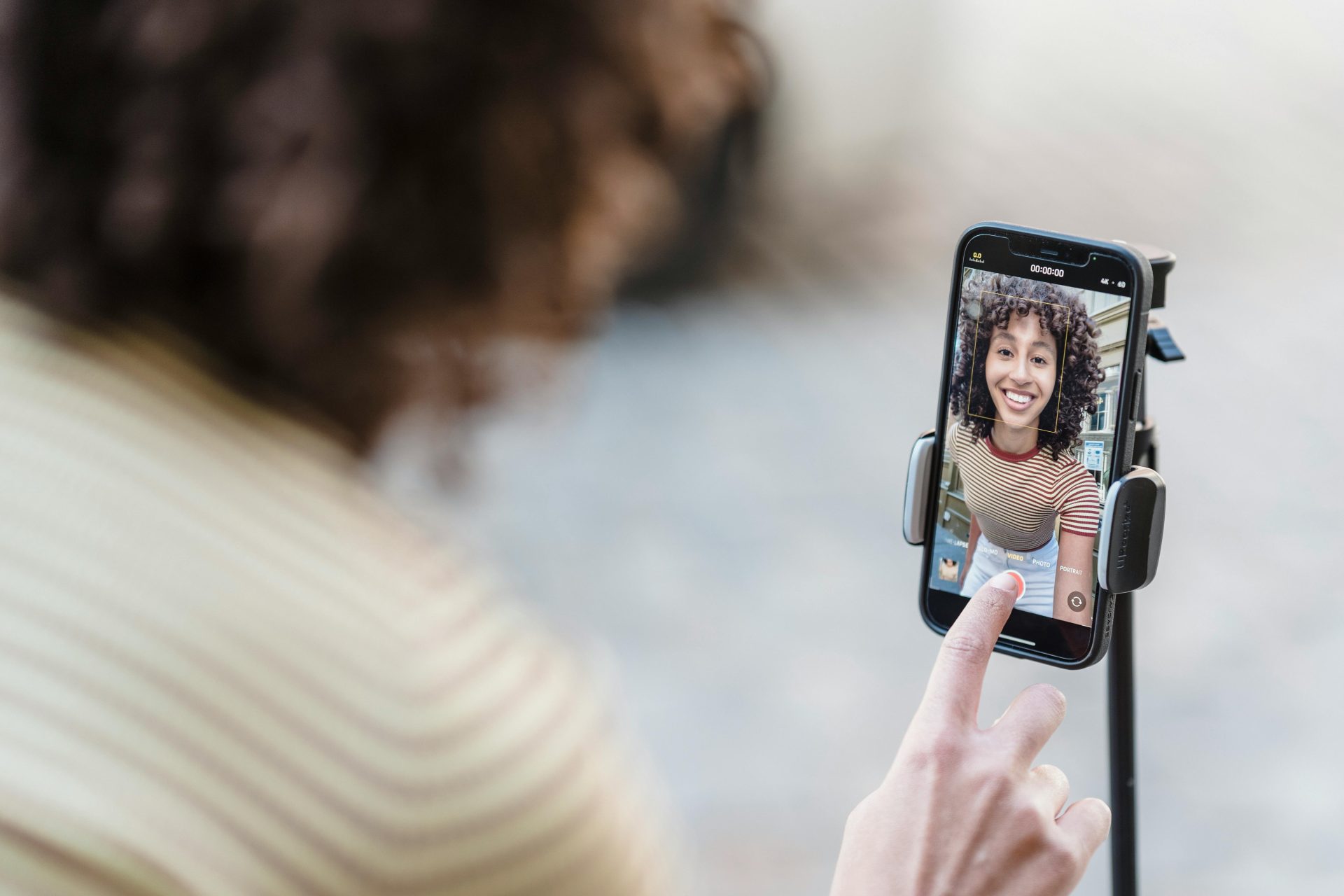 pexels-blue-bird-7243156 A shot from over the right shoulder of a young woman shows her phone on a tripod stand. On the phone screen as it's facing her, you can see her smiling and about to press the record button