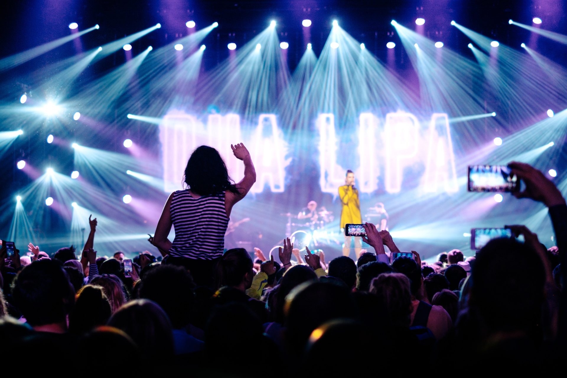 pexels-dua-lipa-1838653 A woman on someone's shoulders with her right arm up in a packed audience all dancing to Dua Lipa. Dua can be seen on stage with 'DUA LIPA' written behind her on screen