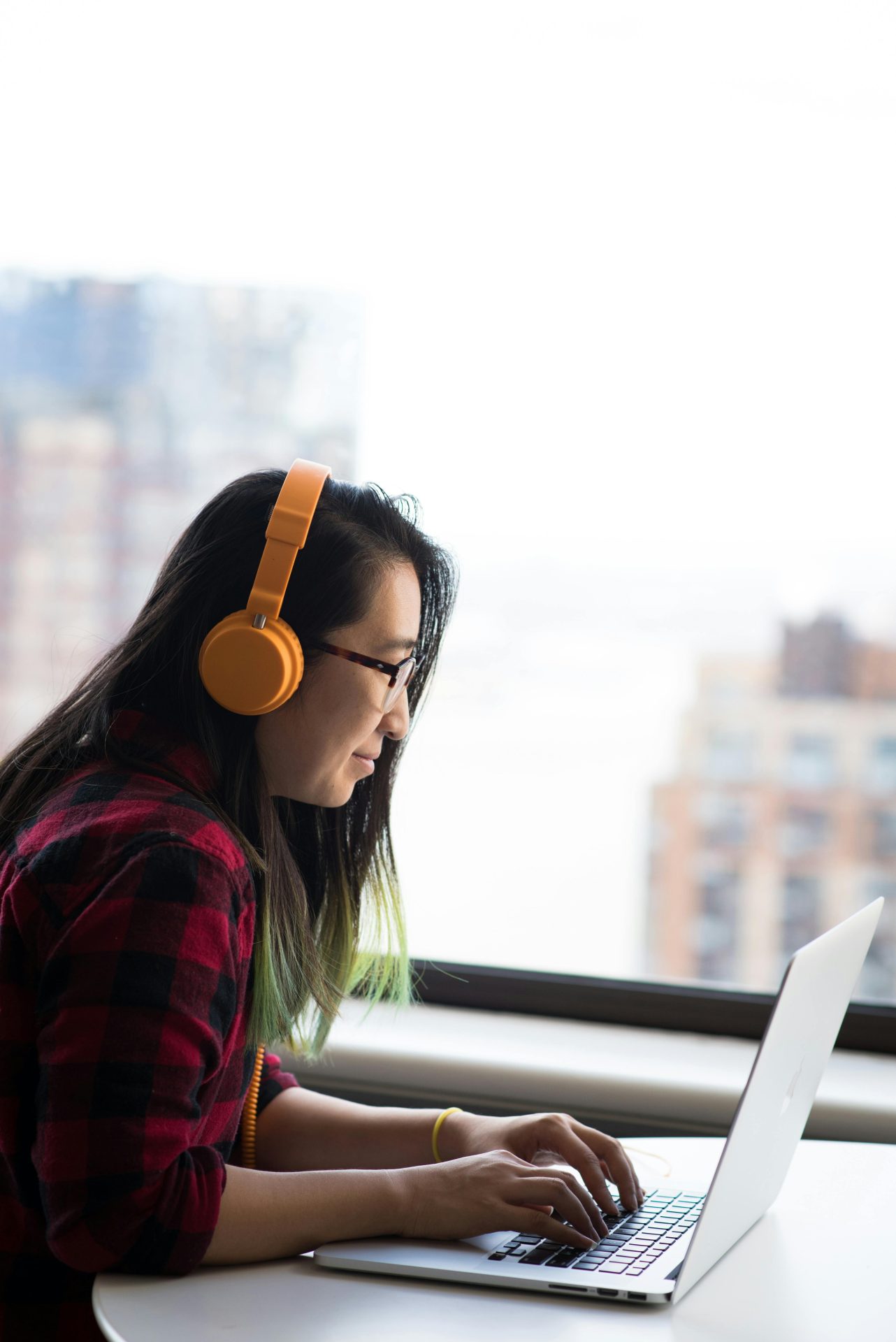 pexels-divinetechygirl-1181682 Portrait image of a woman with dark hair wearing glasses and a check shirt working on her silver laptop wearing orange headphones