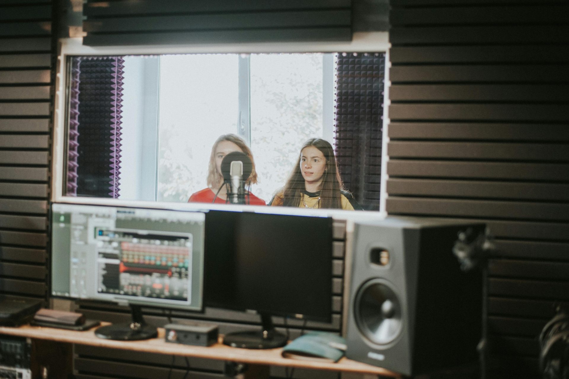 pexels-john-taran-166597215-11044817 Two women pictured through a glass window in a sound booth in front of a microphone