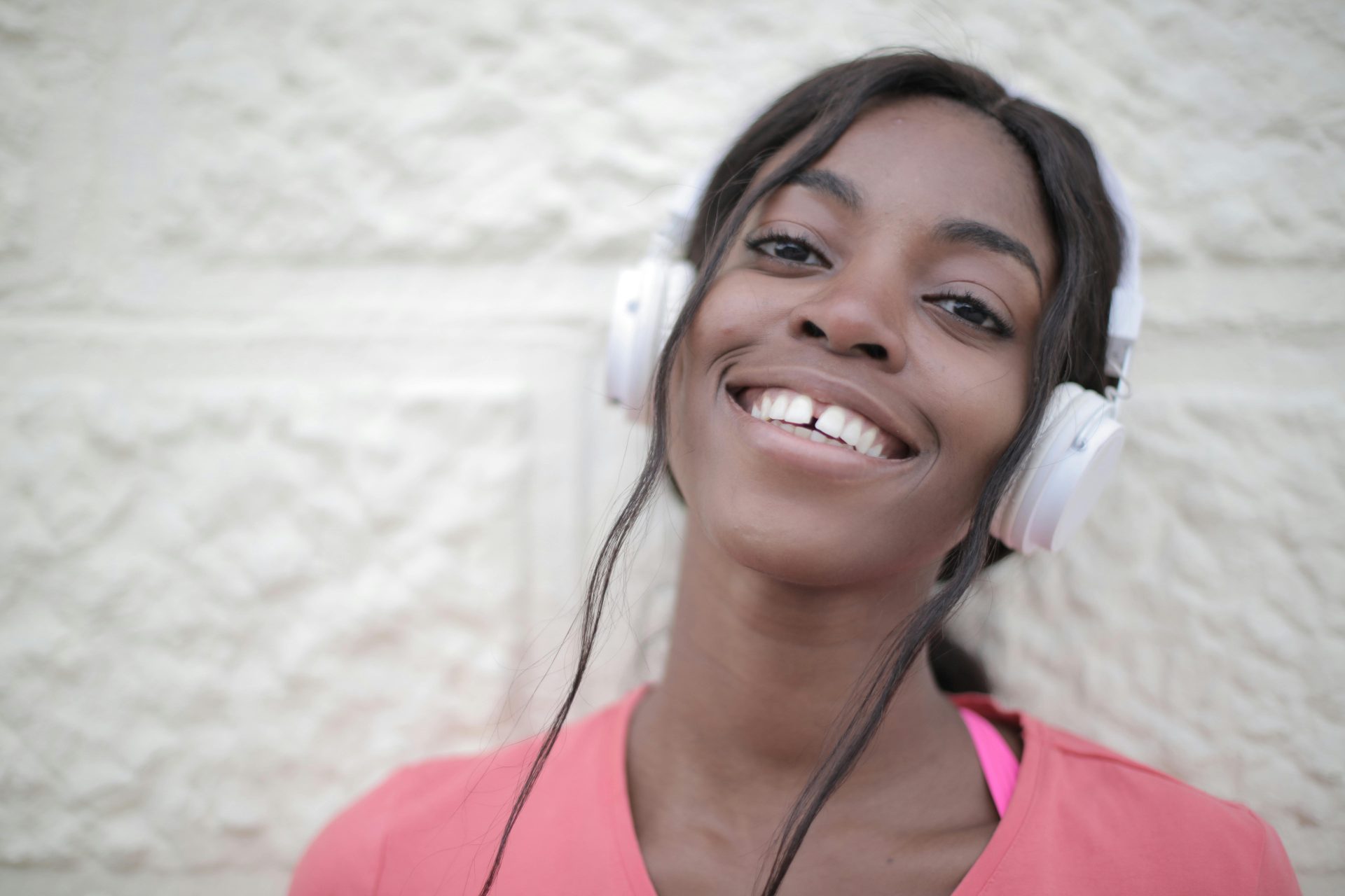 pexels-olly-3800500 A close up of a woman with white headphones on smiling at the camera in front of a white brick wall