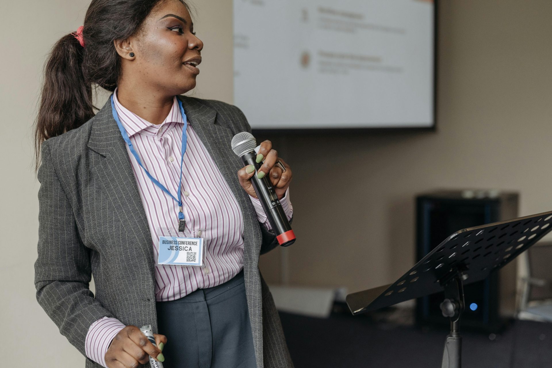 pexels-pavel-danilyuk-8761541 A black woman wearing a suit is holding a microphone stood up in front of a projector and a stand, looking to the right