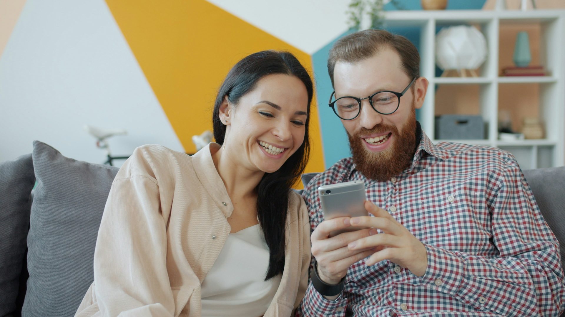 vitaly-gariev-Mq3HfpG5DkM-unsplash A man with glasses, a beard and a check shirt is holding a smart phone and a woman is sat next to him on the sofa. They are both smiling at the phone